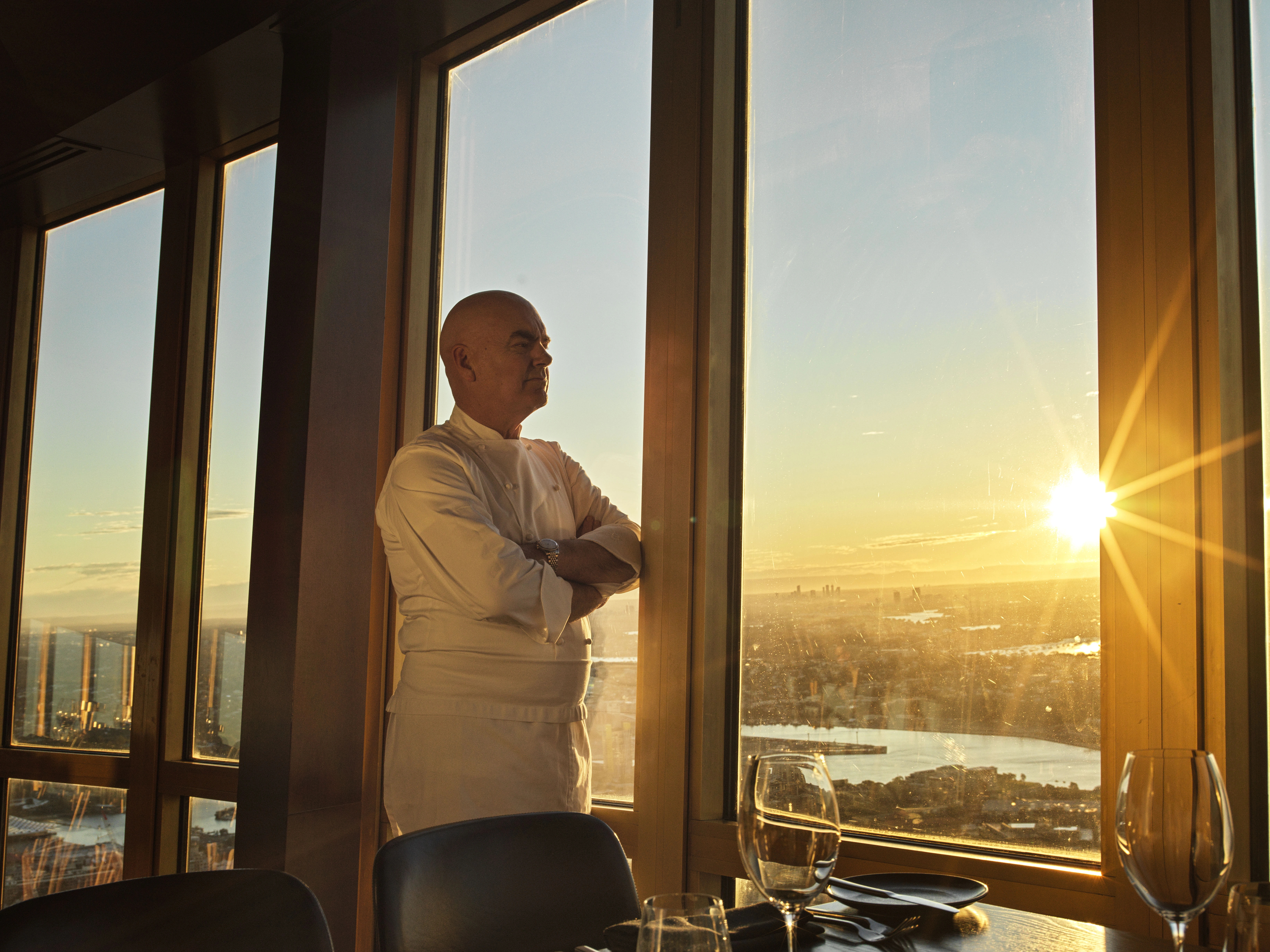 A chef in a white jacket stands by floor-to-ceiling windows at sunset, overlooking a city skyline and water.