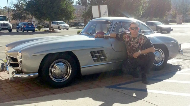 Mark Balelo posing beside a silver classic Mercedes car outdoors in daylight.