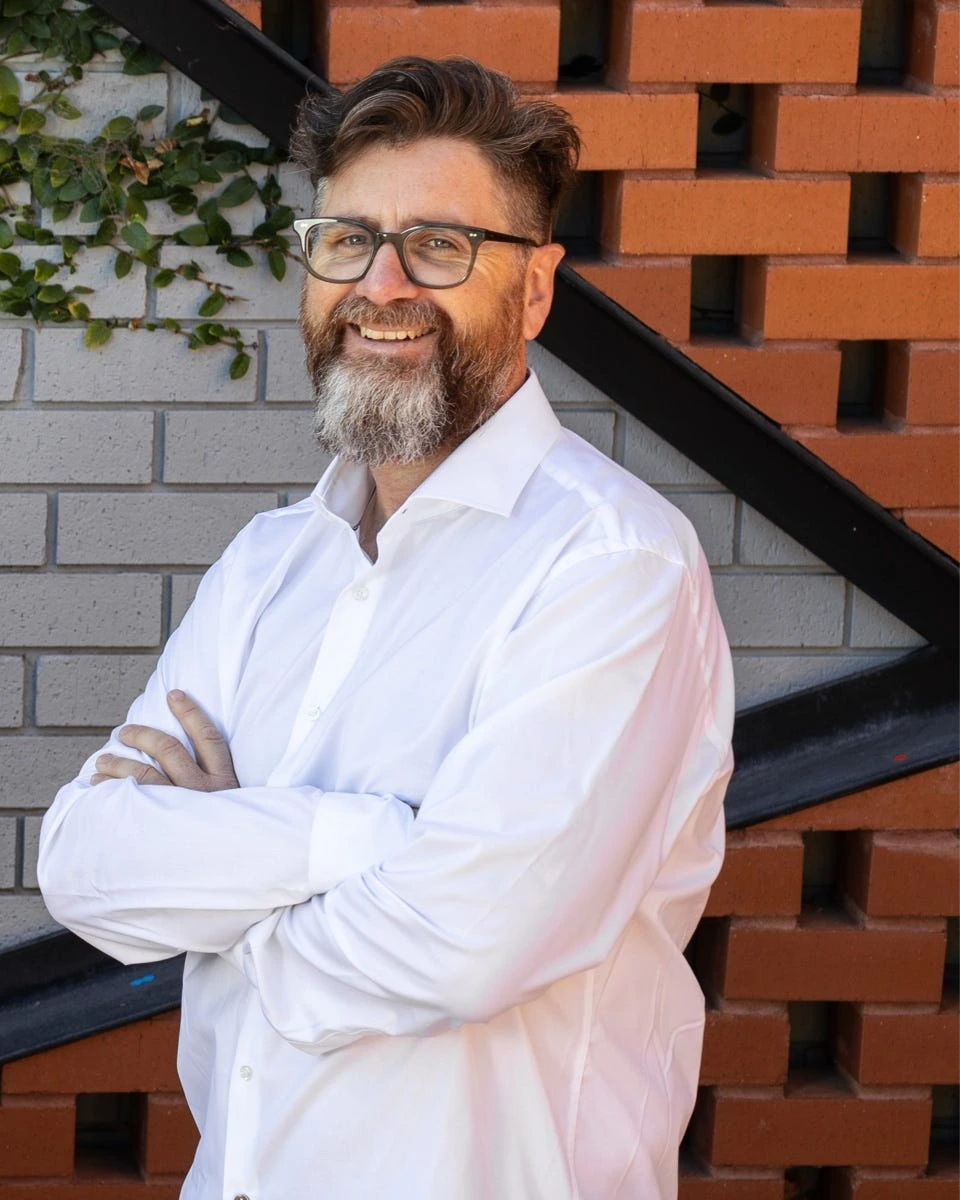 Smiling man with glasses and beard standing against a brick wall